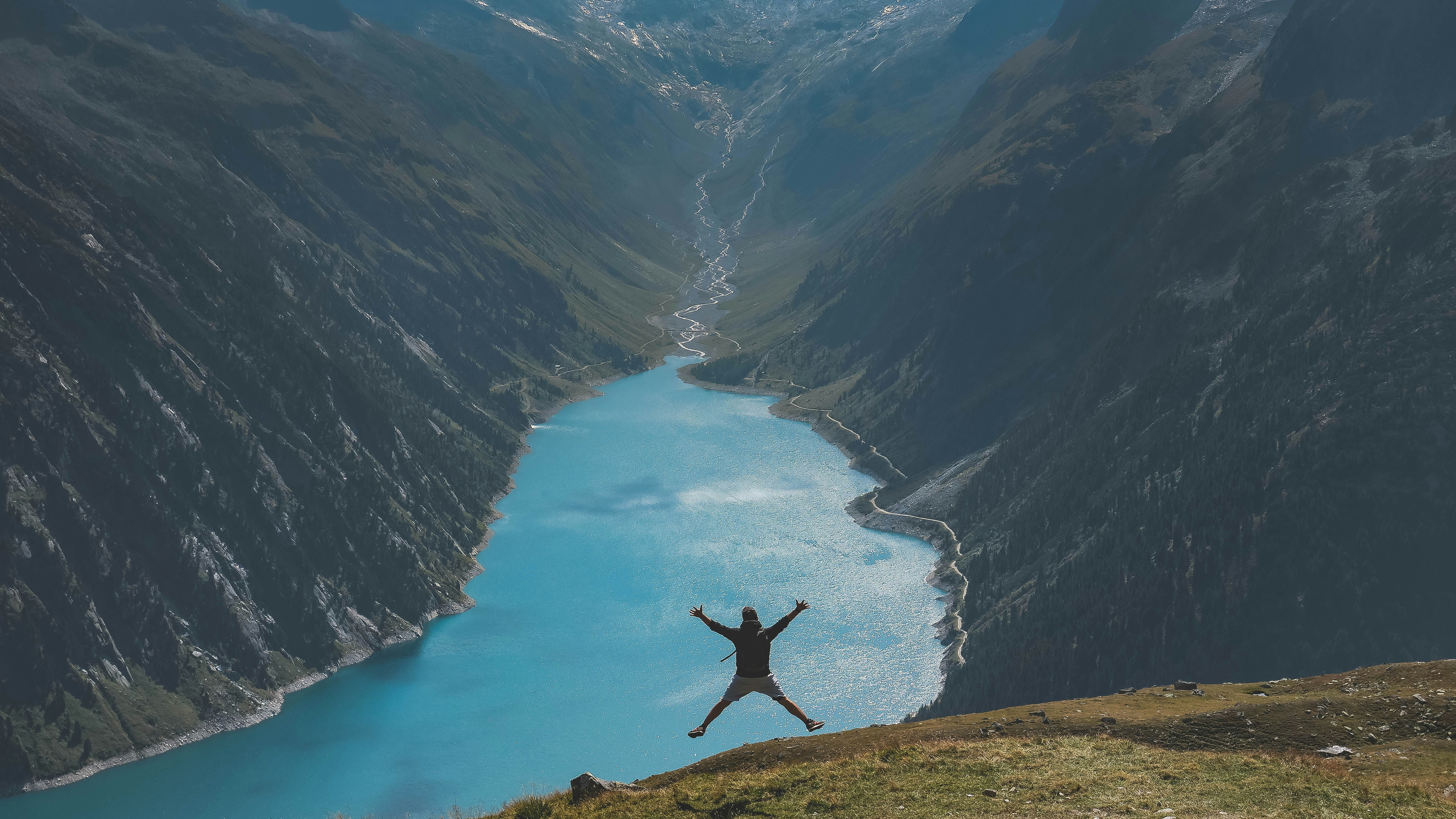 Un homme fait un saut de joie devant un lac au coeur des montages, la photo est prise de loin pour montrer le paysage